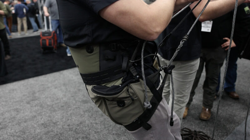 A hunter leans back to demonstrate the olive green and black Arsenal Cloud Freedom hunting saddle, showing the full-support panel, integrated side zipper pockets, and the X2 dual bridge rope system under tension at a trade show.