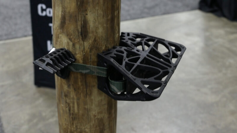 A black, injection-molded nylon Buzzard Roost Pursuit wedge platform and a single Bullman climbing step attached to a wooden post with a camo webbing strap, showing a hybrid ring of steps setup.