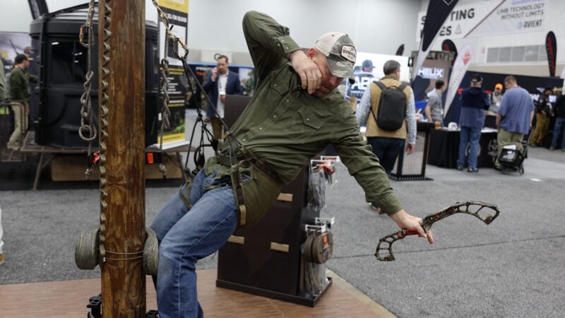 A hunter in a green shirt and camo hat hangs suspended from a Tethrd tree saddle, leaning far out with a mock compound bow while standing on a mobile hunting platform attached to a wooden demonstration post at a trade show.
