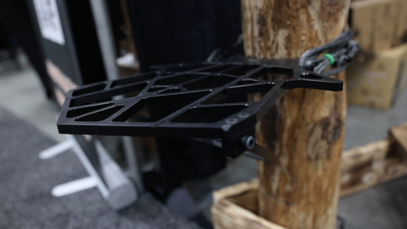 A black Timber Ninja MK Mini-Kunai saddle hunting platform secured to a wooden post, showing the ultra-thin, postless deck design with integrated side wings and a bottom stability spike engaging the bark.