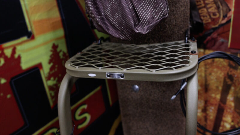 Close-up of the steel mesh top platform on the olive drab Trophy Treestands Hangtime Ladder system, showing the 12 by 9-inch stepping deck mounted against a simulated tree with a product ID tag on the front edge.