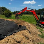 An orange Kubota mini excavator with the brand name visible on the arm sits on a dirt berm next to a large dugout water hole basin lined with black fabric. A green crop field and trees sit under a bright blue sky with white clouds in the background.