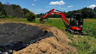 An orange Kubota mini excavator with the brand name visible on the arm sits on a dirt berm next to a large dugout water hole basin lined with black fabric. A green crop field and trees sit under a bright blue sky with white clouds in the background.