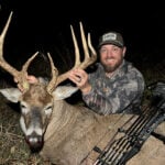 A nighttime hero photograph of bowhunter Justin Zarr smiling behind his harvested 165-inch Kansas whitetail buck in tall grass. Justin wears a camo jacket and a brown ScentLok logo cap while holding the buck's tall antler tines. Resting across the deer's body is a grey camo compound bow loaded with Easton arrows featuring white and pink fletchings.