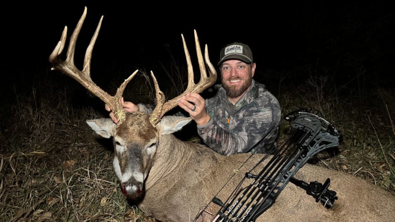 A nighttime hero photograph of bowhunter Justin Zarr smiling behind his harvested 165-inch Kansas whitetail buck in tall grass. Justin wears a camo jacket and a brown ScentLok logo cap while holding the buck's tall antler tines. Resting across the deer's body is a grey camo compound bow loaded with Easton arrows featuring white and pink fletchings.