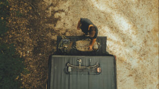 An overhead aerial view of a bowhunter standing at the open tailgate of a pickup truck organizing mobile hunting gear. A compound bow equipped with a quiver of arrows sits on the black tonneau cover, while a saddle platform and tan hunting pack rest on the tailgate along a dirt road.