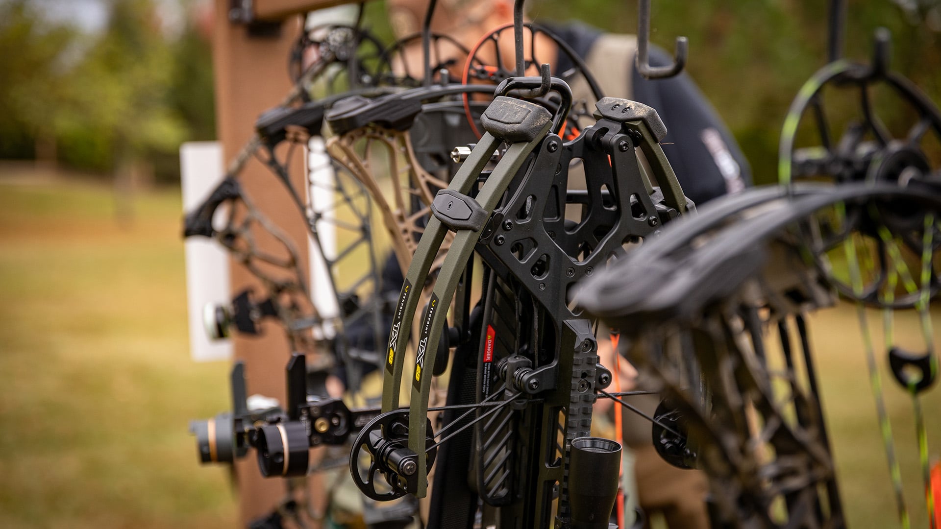 A close-up photograph of an olive drab TenPoint TX crossbow hanging on a wooden outdoor bow rack. Yellow and black text on the crossbow limbs clearly reads TX. In the background and surrounding hooks, several Mathews compound bows hang in a row, visually demonstrating the mix of crossbows and vertical compound bows at an archery range.