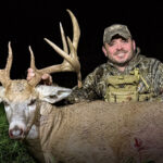 205" Illinois Giant! The Story Of Wags - Smiling hunter in camouflage holding the large antlers of a trophy whitetail buck outdoors at night, with a shield logo featuring an arrowhead and antler graphic in the bottom left corner.