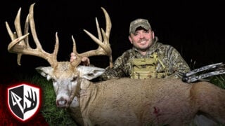 205" Illinois Giant! The Story Of Wags - Smiling hunter in camouflage holding the large antlers of a trophy whitetail buck outdoors at night, with a shield logo featuring an arrowhead and antler graphic in the bottom left corner.