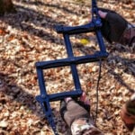 Close-up photograph of a hunter wearing camouflage holding an expanded black B7 Outdoors XP3 Pocket Stick climbing stick. The image highlights the folding metal scissor-linkage mechanism, offset steps, and attached Amsteel rope against a sunlit forest floor covered in autumn leaves.