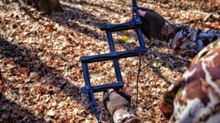 Close-up photograph of a hunter wearing camouflage holding an expanded black B7 Outdoors XP3 Pocket Stick climbing stick. The image highlights the folding metal scissor-linkage mechanism, offset steps, and attached Amsteel rope against a sunlit forest floor covered in autumn leaves.