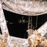 View through a camouflage ground blind window showing an archer inside holding a compound bow with a nocked arrow.