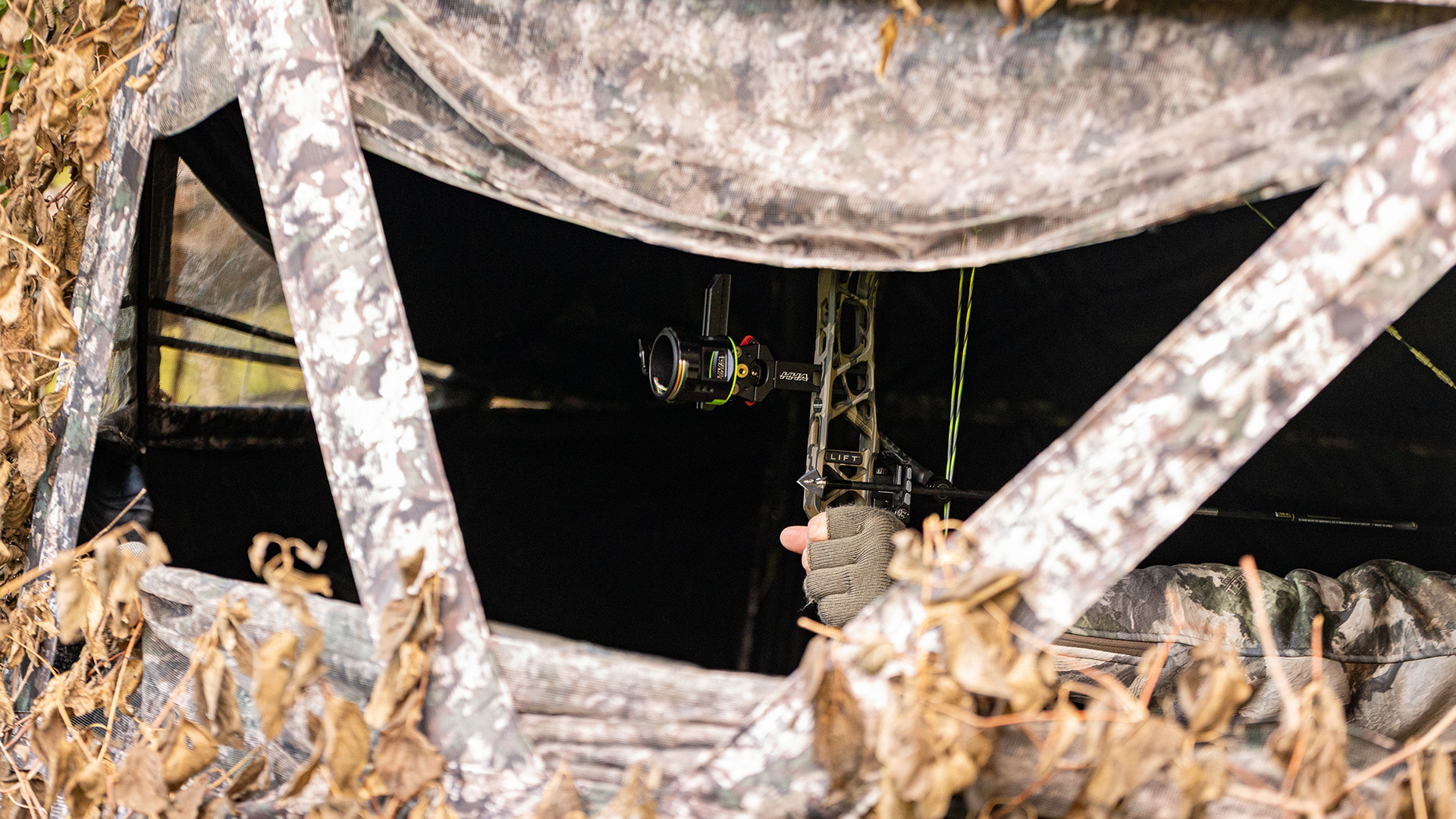 View through a camouflage ground blind window showing an archer inside holding a compound bow with a nocked arrow.